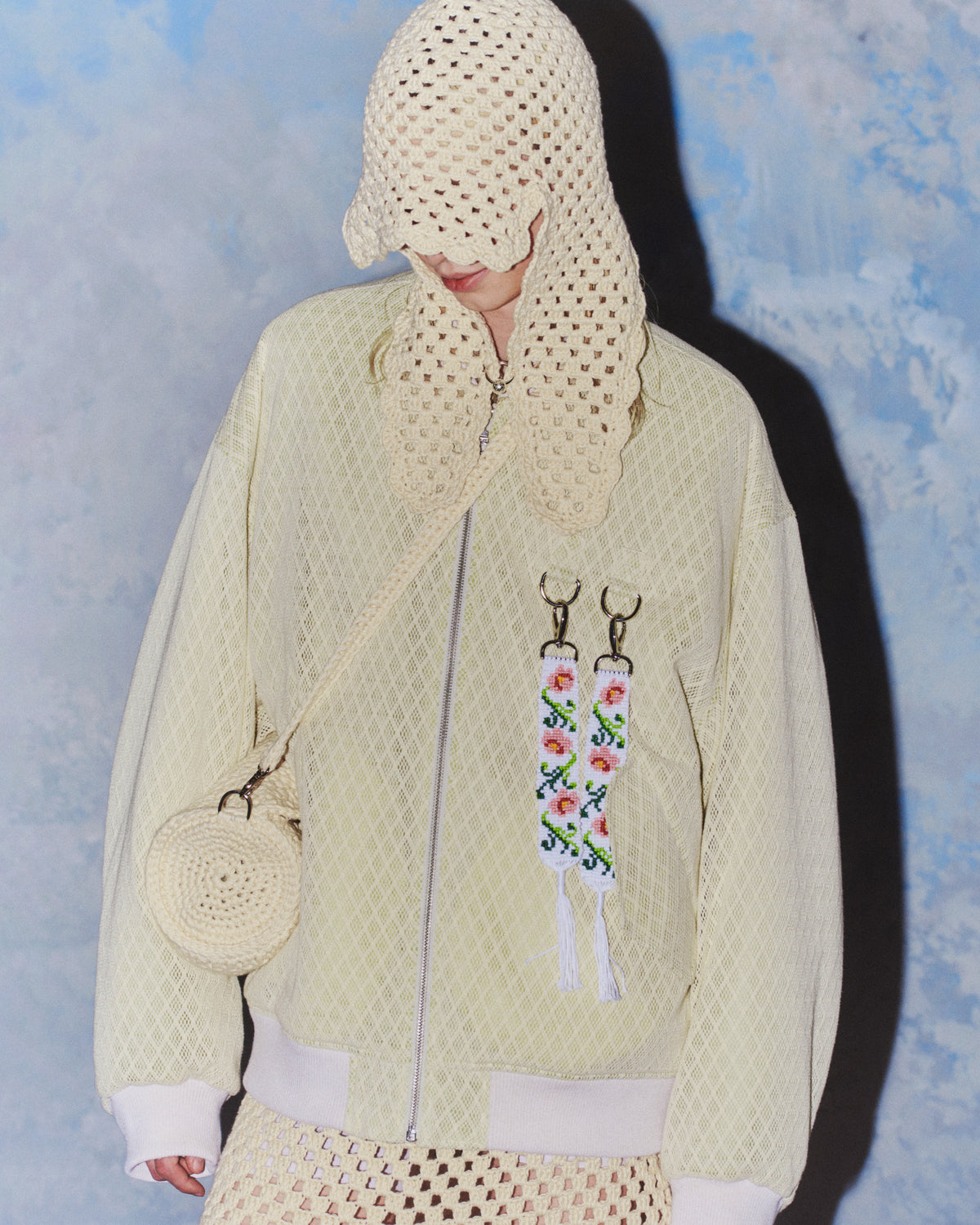 Mallilla on luonnoinvalkoinen takki sekä virkattu mekko, olkalaukku ja huppu, johon on kiinnitetty RAKAS Rolf Ekroth x Kalevala -koru.
